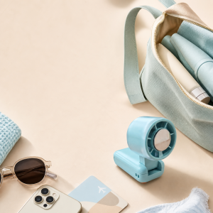 02-yoliton-foldable-handheld-fan-travel Folded Yoliton portable fan beside a travel bag, sunglasses, and phone